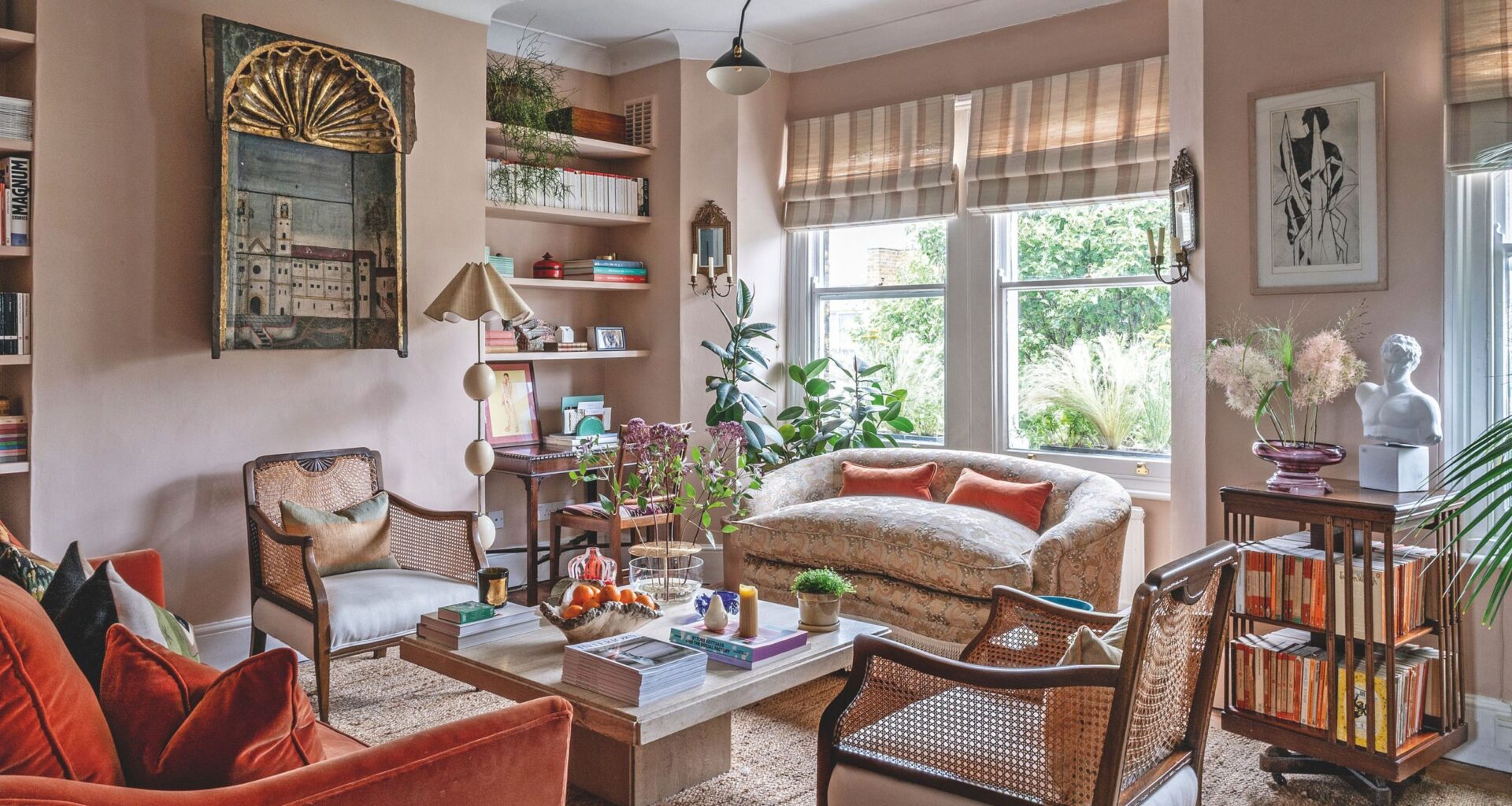 A warm, eclectic living room featuring dusty pink walls, a burnt orange velvet sofa, and woven cane armchairs arranged around a stone coffee table