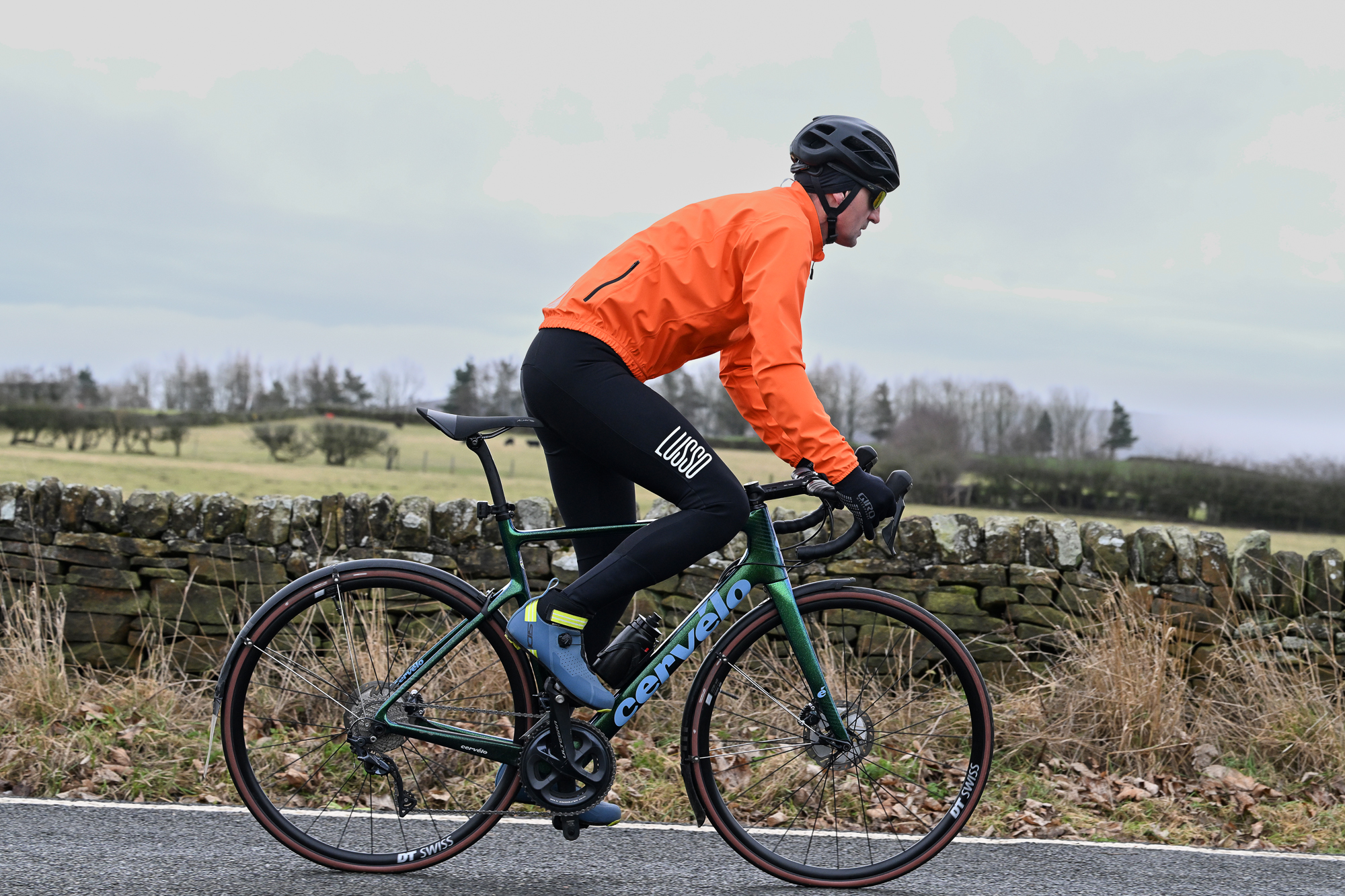 Side view of a man wearing black tights, blue shoes and an orange jacket riding a green road bike