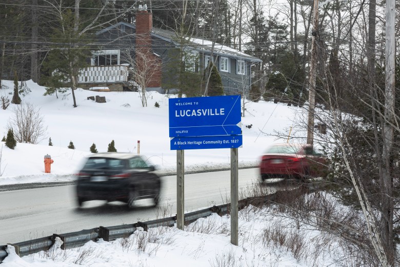 Blurry vehicles are seen on the road near a blue sign reading, WELCOME TO LUCASVILLE, HALIFAX, A Black Heritage Community Est. 1827.
