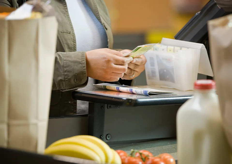 Person paying at a grocery store checkout, holding cash. Items like bananas and milk are visible