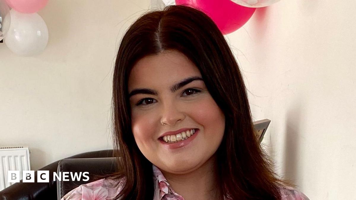 Ayla Jones is a white young woman with shoulder length dark brown hair. She is wearing a floral pink blouse. Behind her on the wall are some clear, white and pink balloons and there is pink confetti on the table. She is smiling directly at the camera.