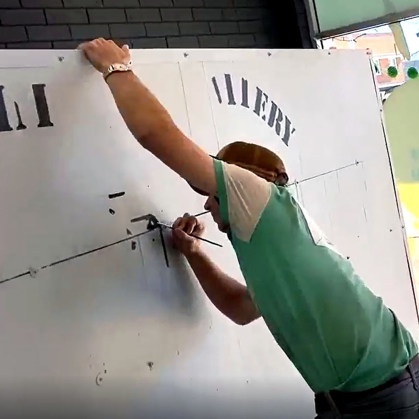 A young man wearing a green t-shirt paints a battery symbol on the side of large grey box taller than he is.