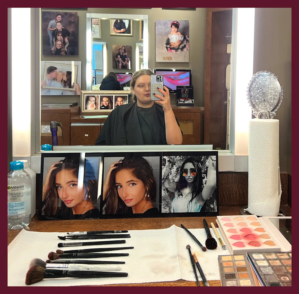Kerry Justich sits in a makeup chair and takes a selfie of her reflection in the mirror. Makeup and framed photos surround her.
