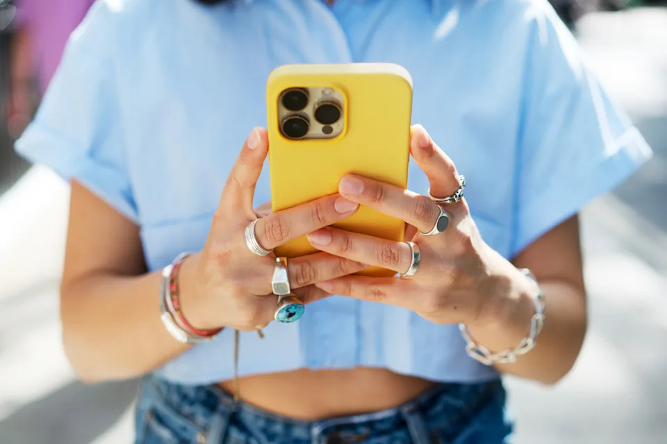 Person outdoors holding a smartphone with a yellow case, wearing a blue shirt, multiple rings, and a bracelet