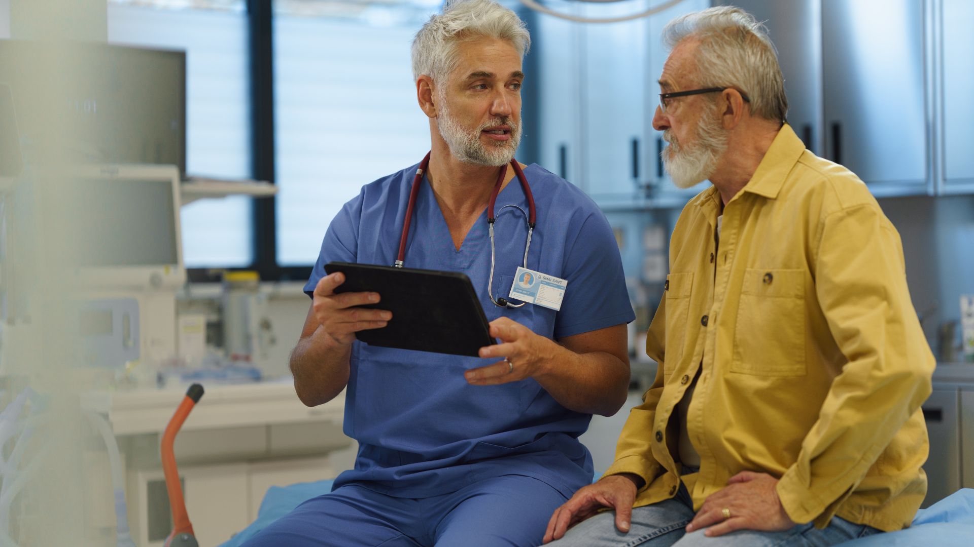 A man aged over 65 with gray hair and beard talks to a medical professional at a hospital.