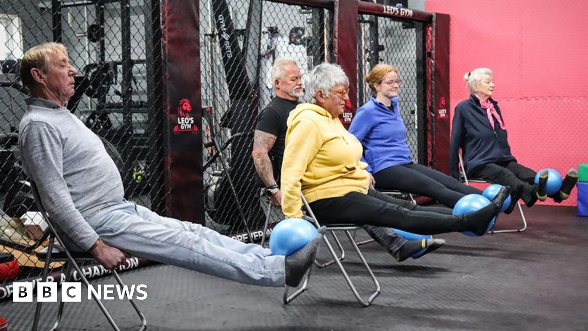 Five members of the class four older and one younger, sit on chairs with their legs outstretched balancing blue balls on their feet