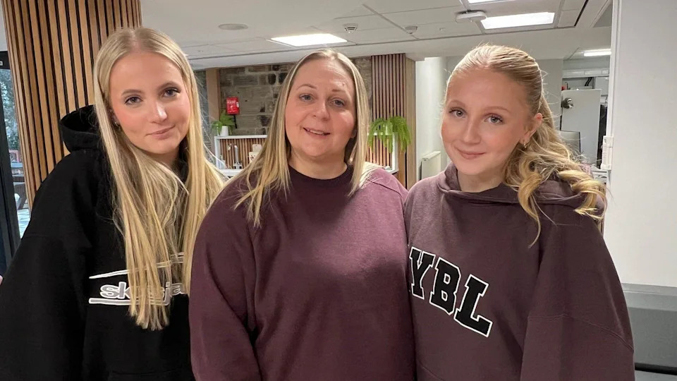 A 45-year-old woman is standing next to her two daughters in the reception area of a university building where the stroke clinic takes places. All three have blonde hair and are smiling.