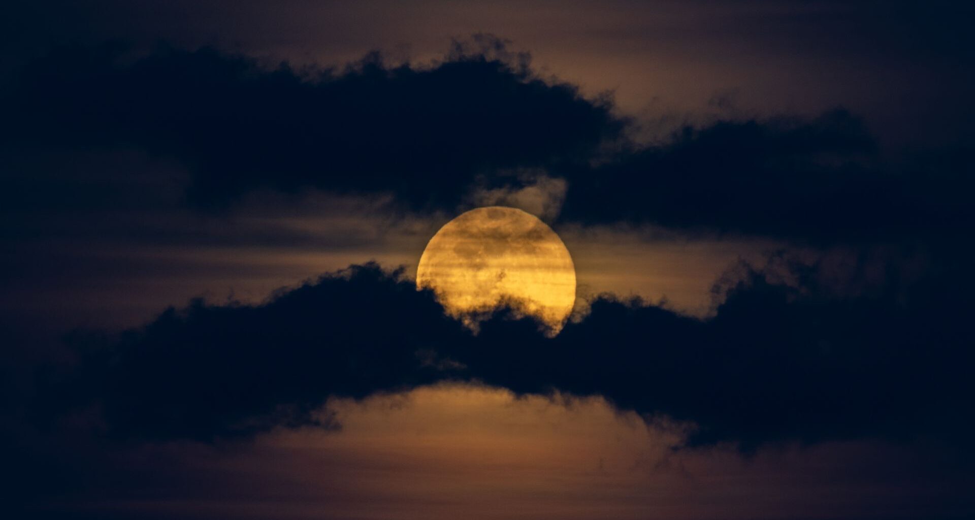 A yellow full moon is pictured glowing in a dark night sky between two worm-like bands of dark clouds.