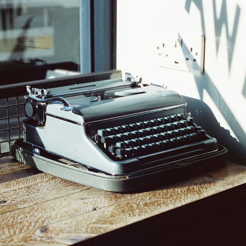 A vintage typewriter sits on a wooden table by a window, illuminated by natural light. A wall socket and shadows are visible on the wall behind it.