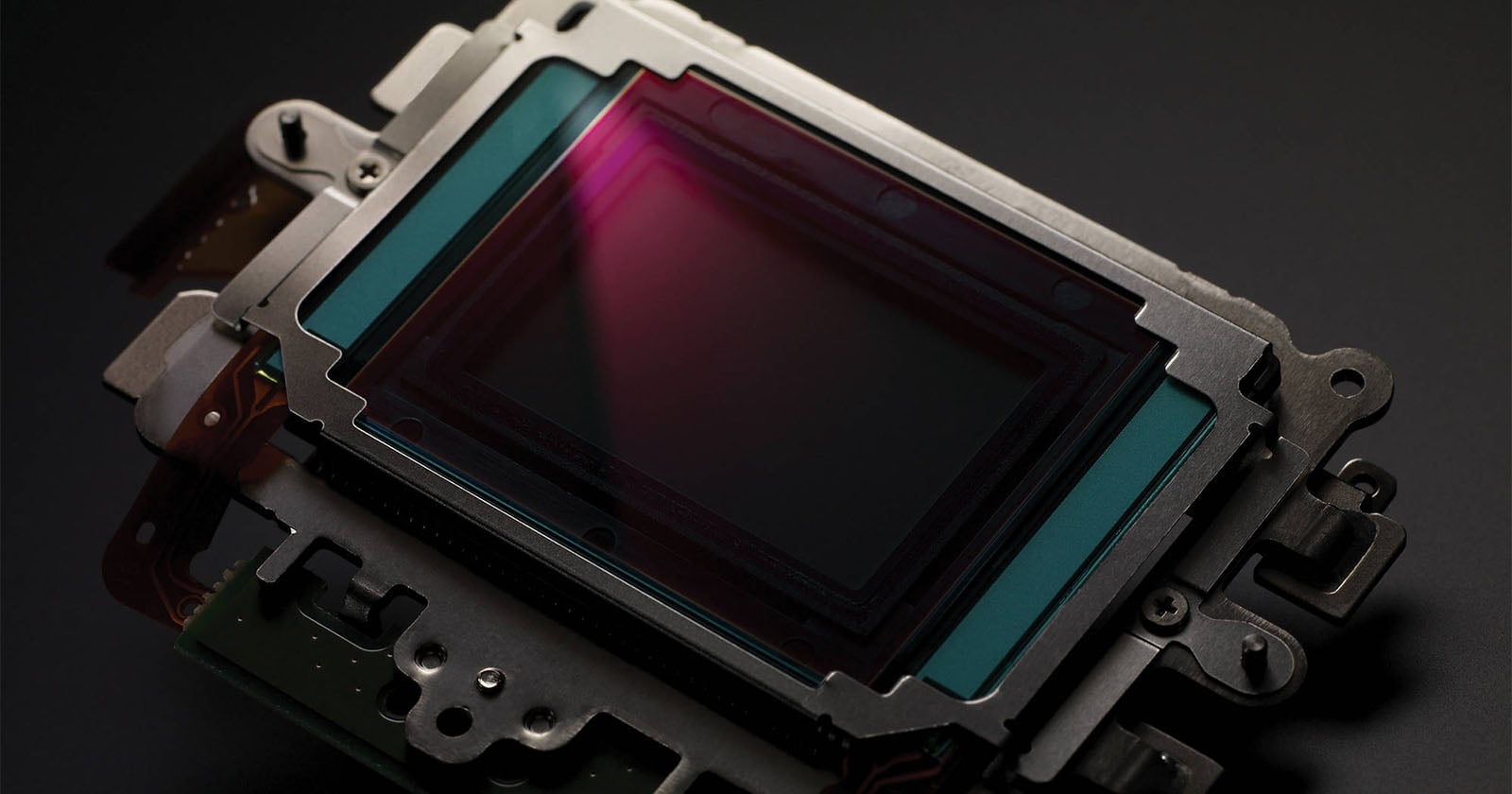 Close-up of a digital camera image sensor on a dark surface, showing its rectangular glass-covered area surrounded by metal and electronic components, with a faint purple reflection on the sensor.