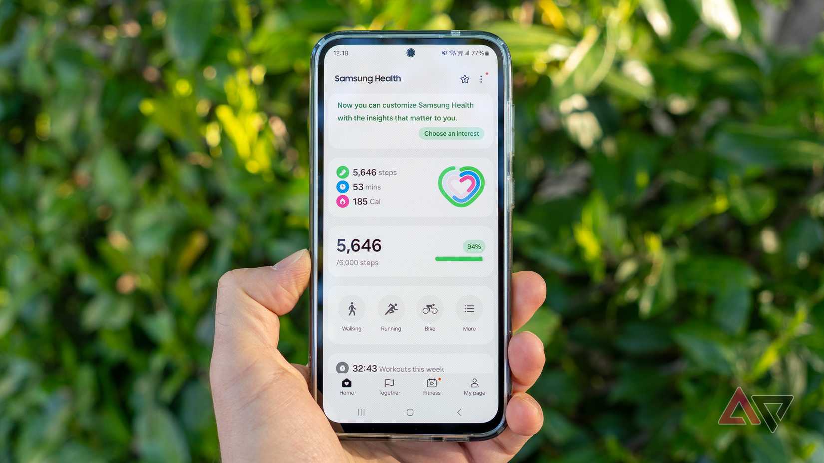 a phone in hand displaying the samsung health app home screen with bushes in the background