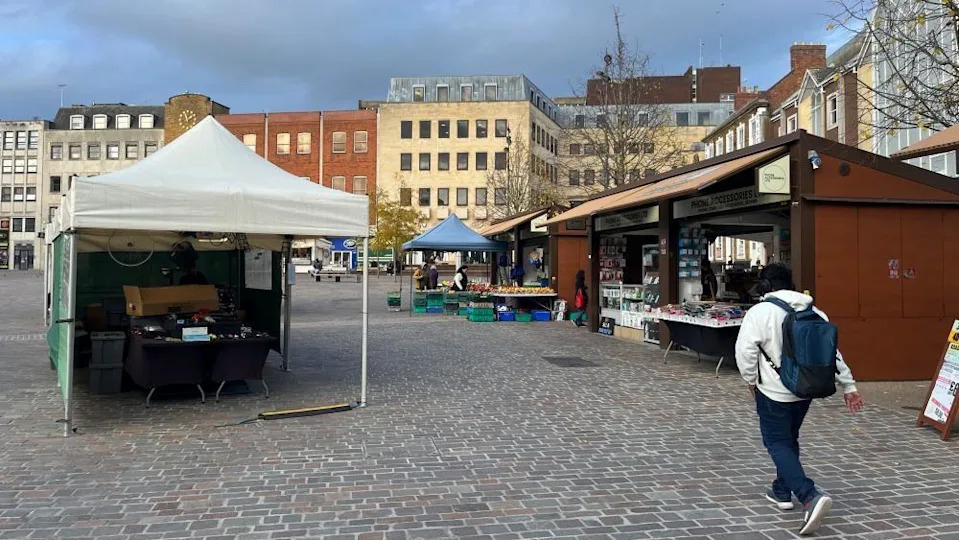 A man, with a white top on, blue trousers, with a blue backpack, walking through a town square, that has cobbles on the ground, a marque, tented stall to the left and back, and static stalls to the right. There are a number of buildings around the square, with trees in the distance. It also shows a blue sky. 
