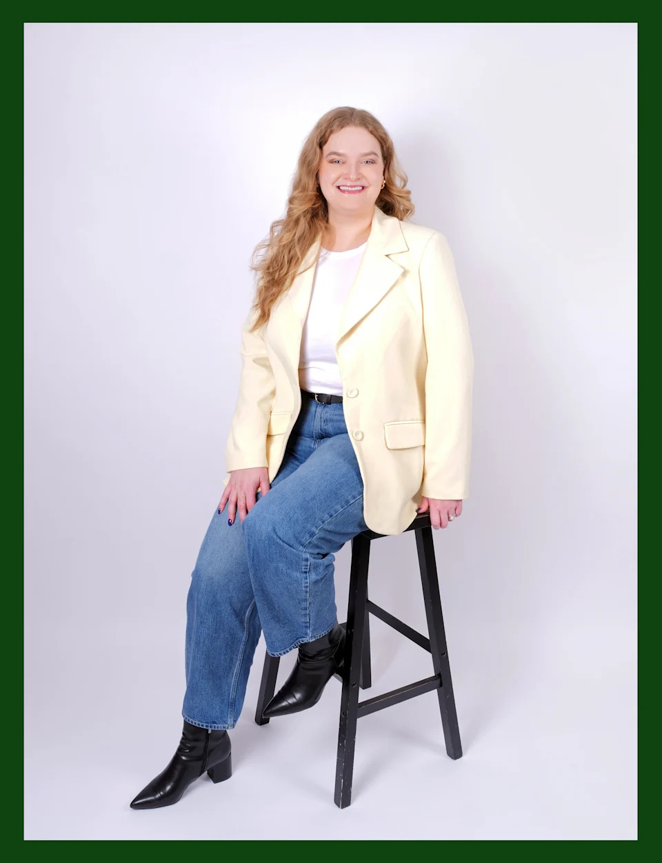 Kerry Justich, in white jacket, jeans and black boots, sits on a wooden stool and poses for a full-length portrait.