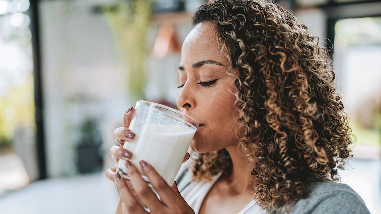 Woman with curly hair drinking a glass of kefir in a bright kitchen setting.