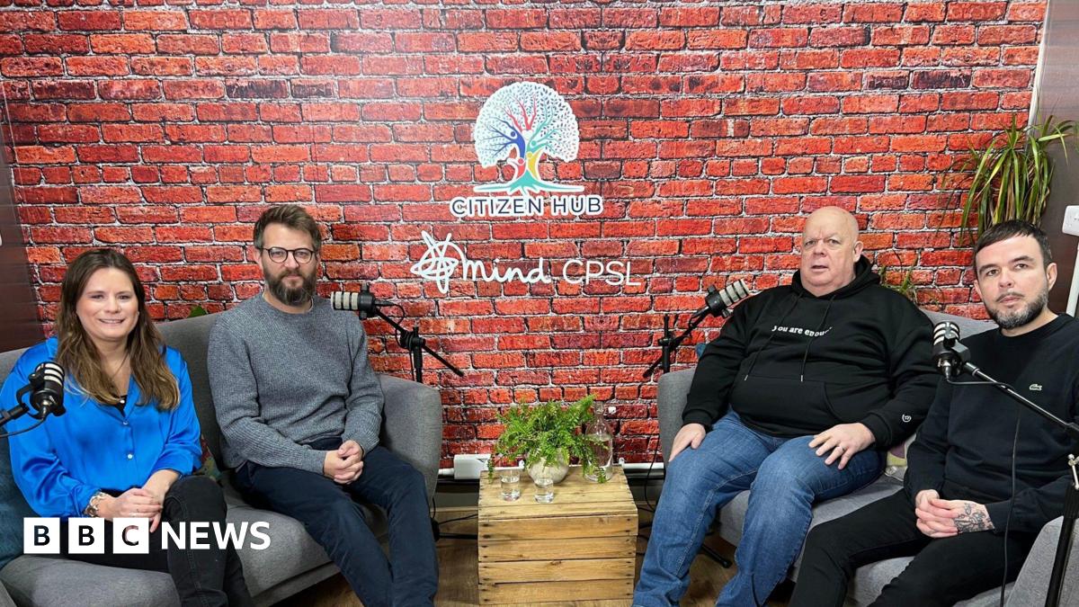 Members of the CPSL podcast team- a woman in a blue blouse, a man in a grey jumper and navy trousers, a man in a black hoodie and blue jeans and a man in a black jumper and black trousers- sitting on grey sofas on either side of the room with microphones in front of them.