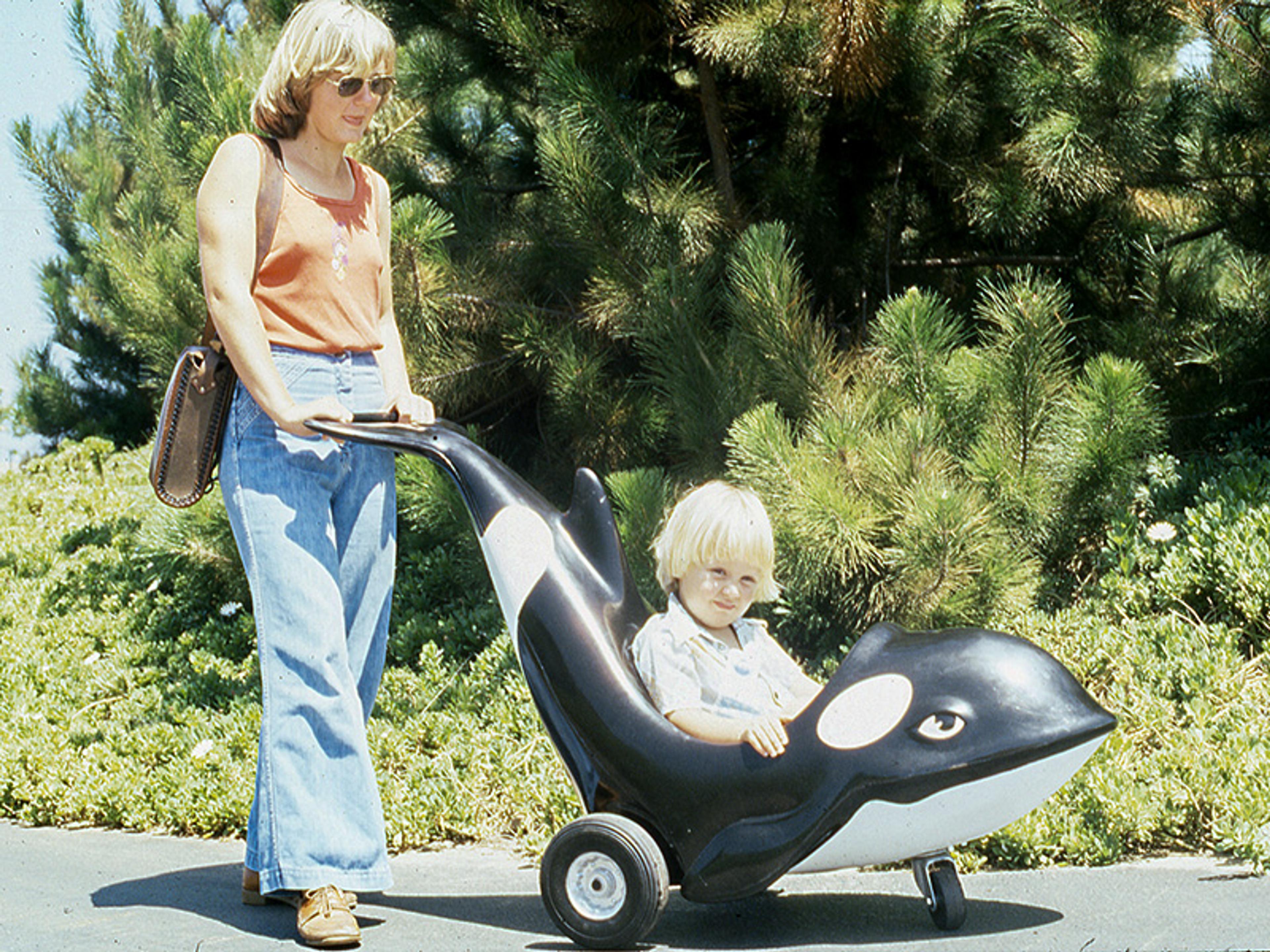 A woman pushing a child in a toy orca cart on a path with greenery and trees in the background.