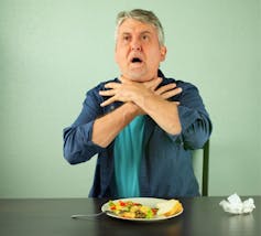 A man sitting at a table choking with his hands at his neck and a plate in front of him