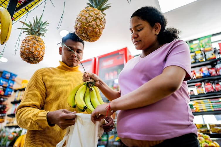 A pregnant woman and a man place a bunch of bananas into a bag while shopping in a grocery store.