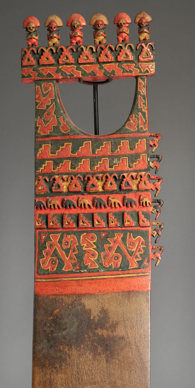 Carved wooden paddle decorated with red, green, and yellow paint, featuring a line of small figures at the top and animal carvings down the center.