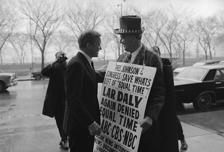 A man holding a placard and wearing a hat speaks for another man in a black and white photo.