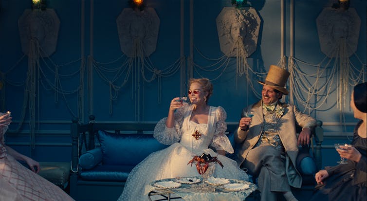 Film still: A woman and man sit in a lavish blue room adorned with Medusa heads and pearls, smiling and laughing while holding drinks.