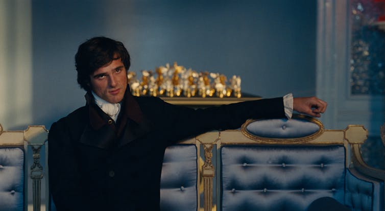 Film still: A brooding man wearing a Georgian-style three-piece suit, with his arm outstretched on an antique sofa.