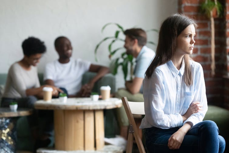 A woman sitting on her own while a group of people her own age chat happily in the background.