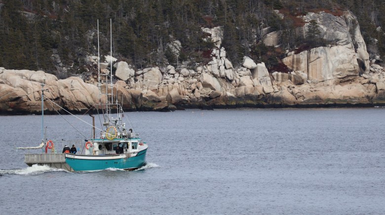 A small fishing boat with a teal green hull and two men on board sails away in a small cove. The shoreline is rock cliffs and forests.