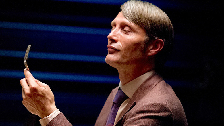 Hannibal Lecter looks down at his fork while wearing a brown suit and blue tie in 