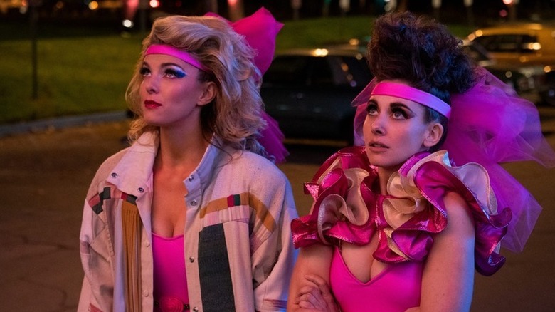 Betty Gilpin as Debbie standing next to Alison Brie as Ruth in pink '80s outfits in GLOW