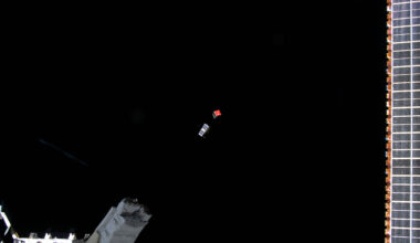 Two CubeSats, one silver and one with a red face, are ejected from the International Space Station (bottom left) into the blackness of space.