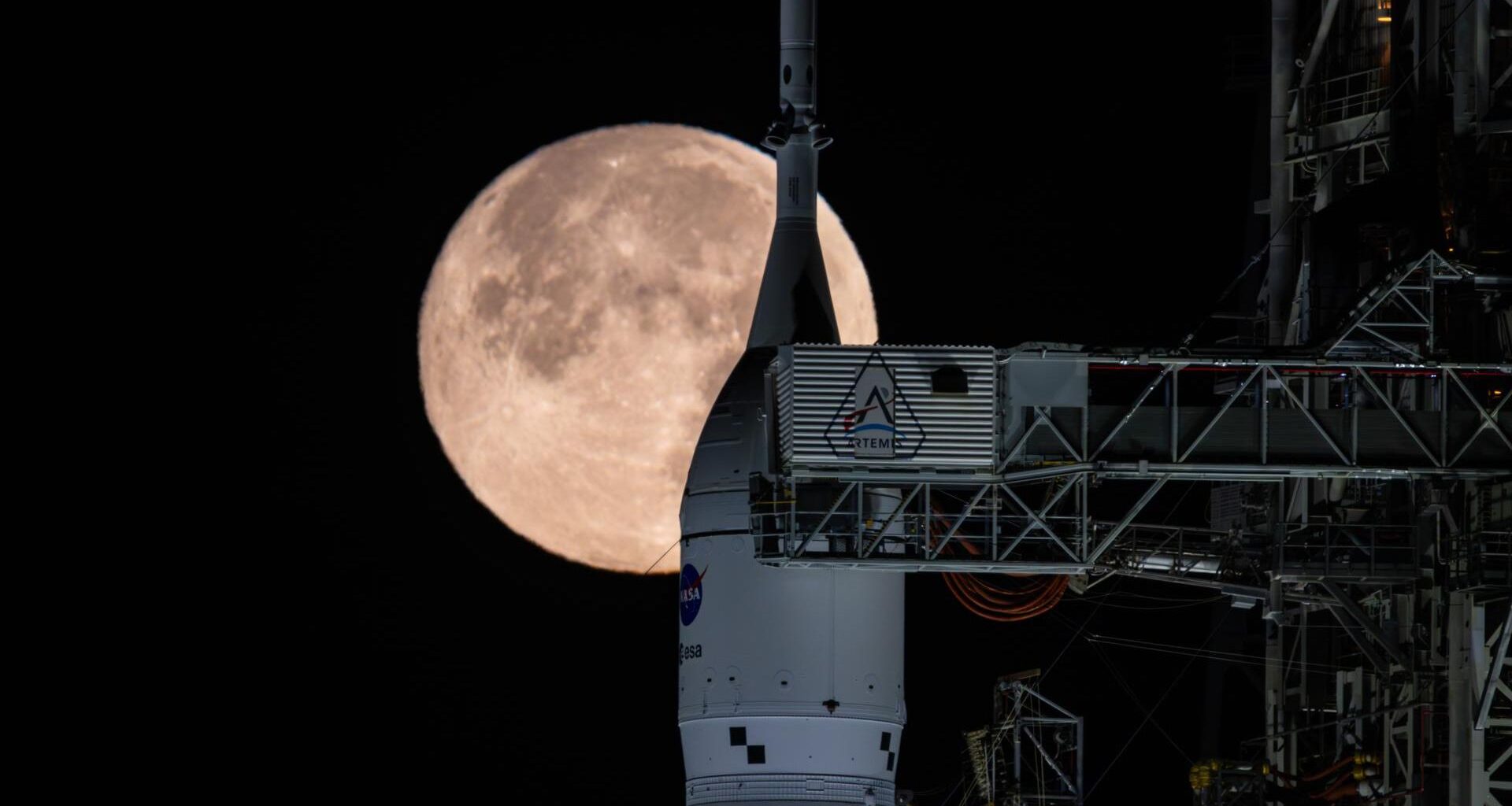 A white rocket nose with the circular NASA logo stands next to the metal gantry underneath a glowing large full moon in a black night sky