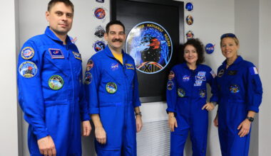 Four people - two men on the left and two women on the right - pose with the Crew-12 mission insignia. They are all wearing blue jumpsuits with various patches on them. The insignia is on the wall, framed in a black recess. Autographed patches are stuck on the wall around the black frame.