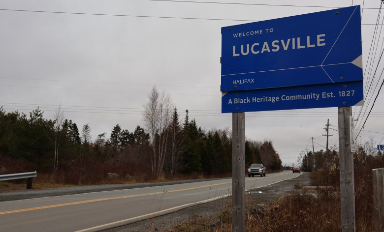 A blue road sign with white text that says "Welcome to Lucasville, Halifax, a Black Heritage Community Established in 1827.