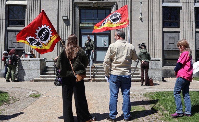 A man and a woman each other a red PSAC flag while standing on a paved pathway in front of an old stone building. A white man is standing on the steps of the building speaking to a crowd. At right is a white woman with longish red hair and wearing a pink T-shirt and jeans.