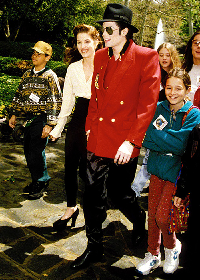 Michael Jackson walking outdoors with children and adults, wearing a red jacket and black hat in a sunny park setting. Michael Jackson walking outdoors with children and adults, wearing a red jacket and black hat in a sunny park setting.