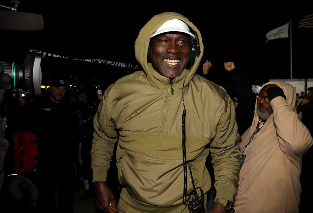 Michael Jordan in a hooded jacket and white cap smiling after Tyler Reddick's NASCAR Cup Series victory.
