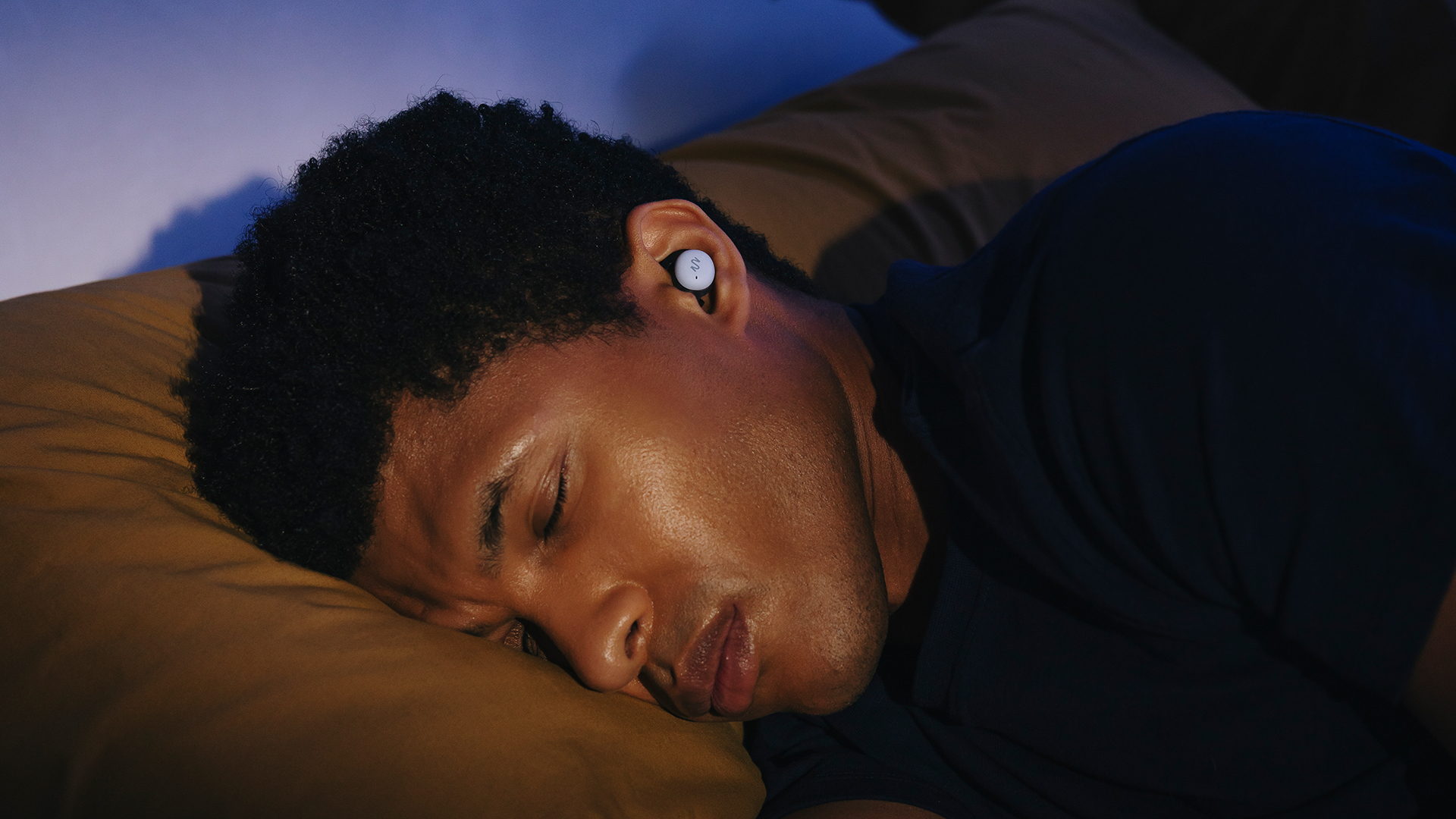 A close up of a man lying in bed asleep on his side in a dark room, wearing a pair of NextSense Smartbuds