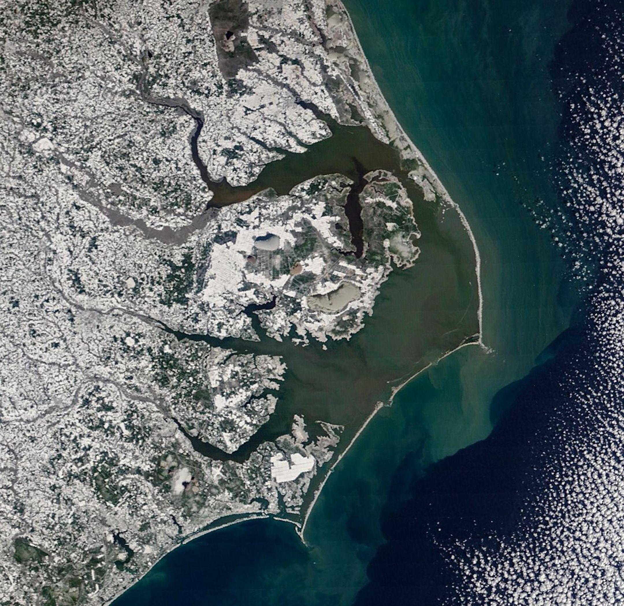 A detailed satellite image centered on eastern North Carolina shows white snow covering coastal areas along the blue-green Atlantic Ocean. Gray ice is visible on some rivers and lakes.