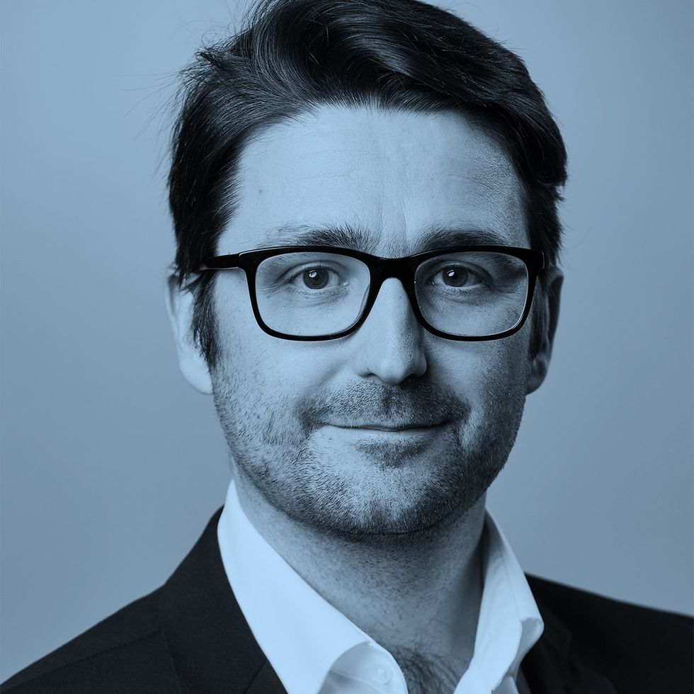 Person in a suit with dark hair against a blue background.