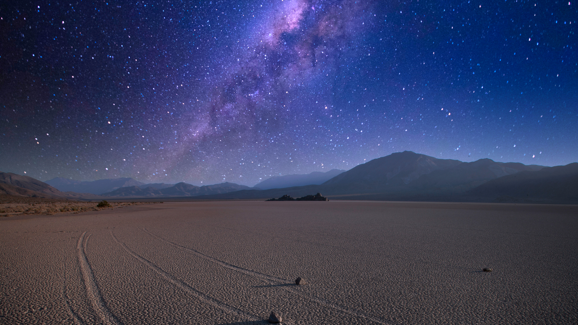 A streak of the Milky Way galaxy glows in a dark blue night sky overlooking a barren desert with a jagged hillside in the background