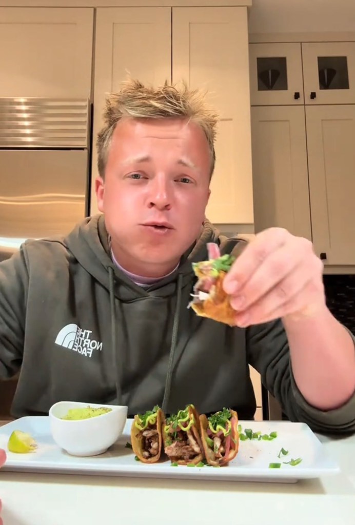 A man wearing a North Face hoodie holds a half-eaten taco while looking at the camera.