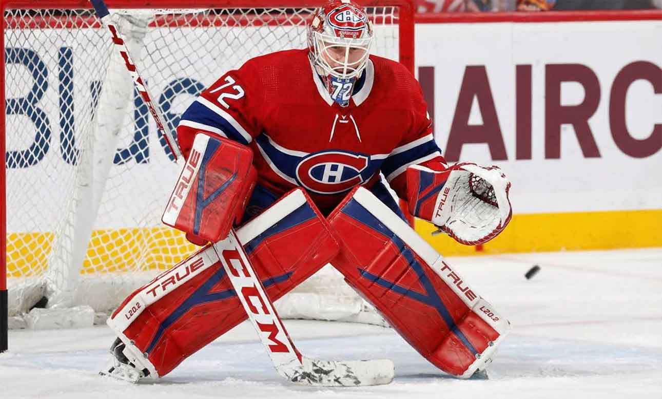 NHL goaltender Sergei Bobrovsky, wearing a red, white, and blue Montreal Canadiens uniform, pads, and mask, is in a butterfly stance on the ice in front of the net. His eyes are focused on a puck sliding on the ice to his right.