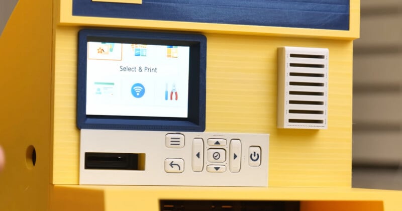 Close-up of a yellow machine with a small screen displaying "Select & Print," several control buttons, a card slot, and a speaker grille. The design has blue and white accents.