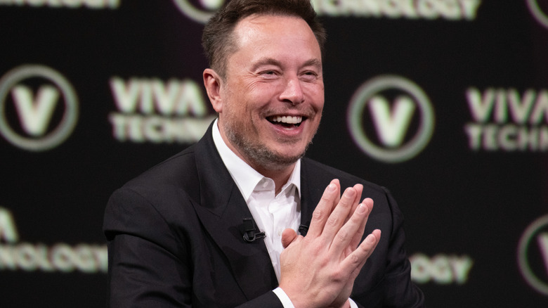 Elon Musk seen at a conference, his hands are held together and he's smiling