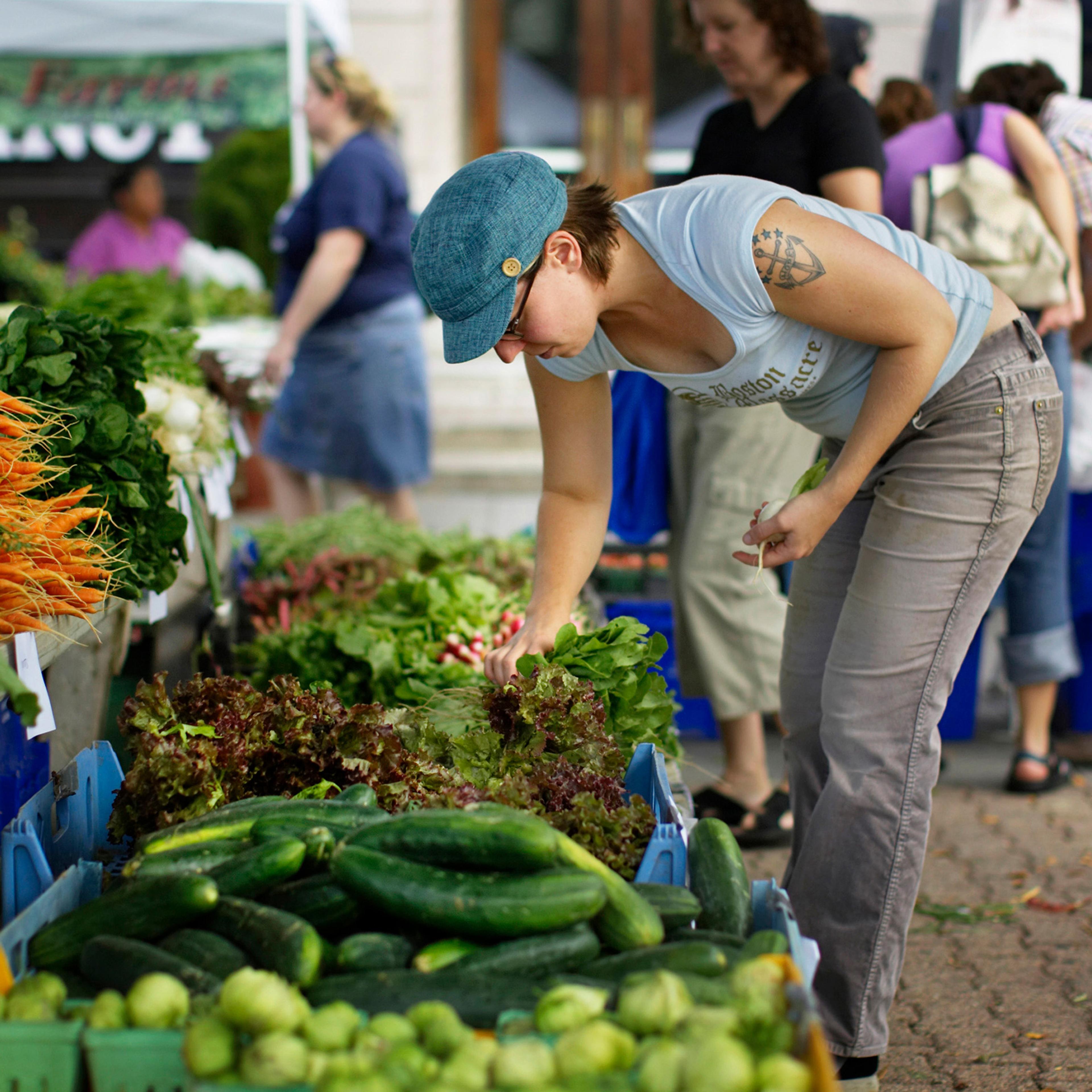 A woman selecting vegetables at an outdoor market stall with assorted greens and carrots available for sale.