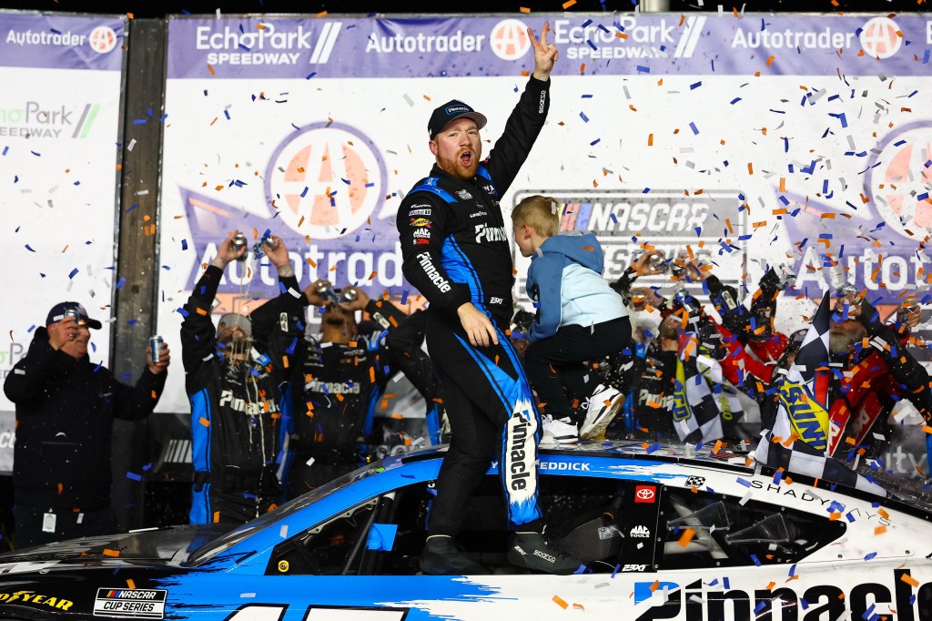 Tyler Reddick celebrates on top of his car after winning a NASCAR Cup Series race, surrounded by confetti and cheering people.