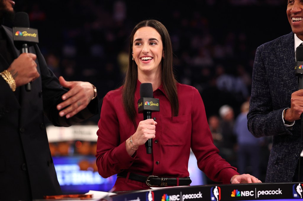 Caitlin Clark smiles while holding a microphone with an NBC Sports logo, on a stage with other people, speaking at an event.