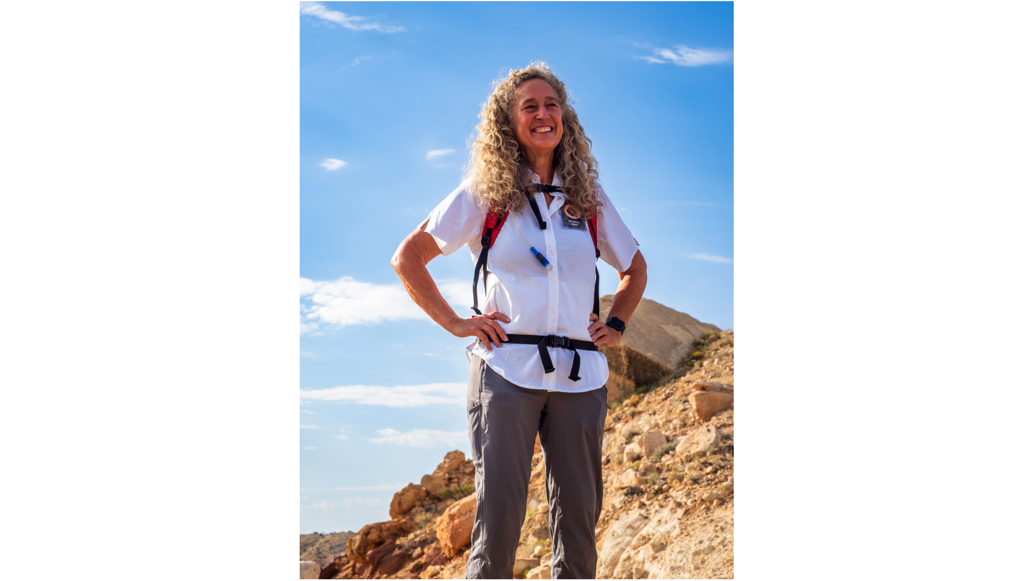 an older woman stands, smiling, on a rocky desert slope