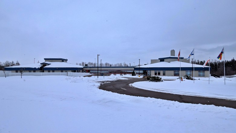 A large industrial looking one-storey beige building is seen on a snow covered landscape, a dark pathway visible through the snow where it was shovelled. Several flags on poles are seen to the right of the photo frame.